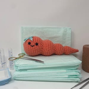 Könnte beinhalten: Ein gehäkelter, korallenfarbener Wurm mit blauer Schleife und lächelndem Gesicht liegt auf hellgrünen Pads. Chirurgische Instrumente, Spritzen und braunes medizinisches Klebeband sind ebenfalls vorhanden, was auf ein medizinisches oder neuartiges Thema hindeutet.