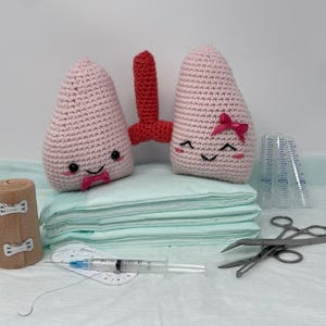 May include: Two crocheted pink lungs with smiling faces, one with a pink bow, on a stack of light blue medical pads. There is a brown bandage, a syringe, and two surgical forceps on the surface.