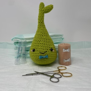 May include: A green crocheted pear-shaped plush toy with a blue bow tie and a smiling face. The toy is sitting on a white surface with a roll of gauze and a pair of surgical scissors.
