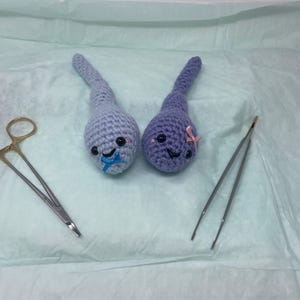 May include: Two handmade crochet sperm cell plushies, one light blue and one purple, each with a smiling face and a small bow. Surgical scissors and tweezers are also in the image. The items are on a light green surface.