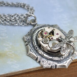 May include: Silver necklace with a detailed pendant featuring a dragonfly perched on a vintage watch face with exposed gears and pink jewels.