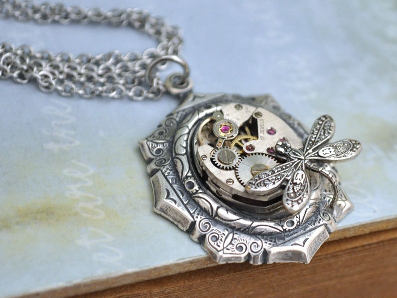 May include: Silver necklace with a detailed pendant featuring a dragonfly perched on a vintage watch face with exposed gears and pink jewels.