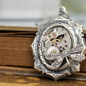 May include: Silver pendant with an intricate design featuring a dragonfly and a vintage watch movement with 17 jewels. The pendant is set against a brown background.