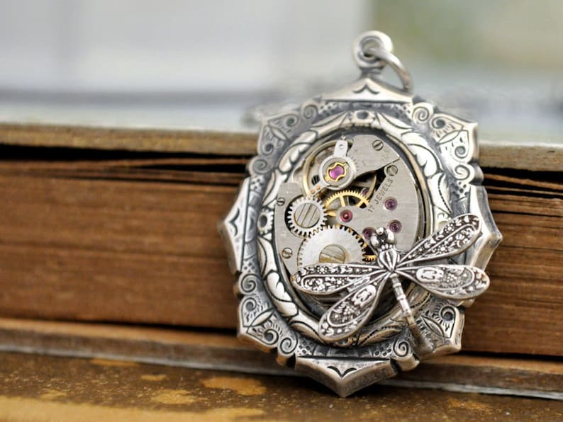 May include: Silver pendant with an intricate design featuring a dragonfly and a vintage watch movement with 17 jewels. The pendant is set against a brown background.