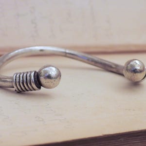 May include: A silver-toned cuff bracelet with a curved design. The bracelet features a ball at each end, with a series of coiled metal rings near one ball. The jewelry is displayed on a light-colored surface.