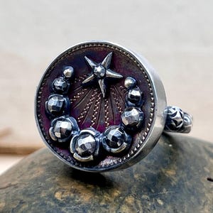 May include: A silver ring with a circular design featuring a central star and surrounding faceted spheres. The ring has a dark purple and silver colour scheme, with intricate detailing.
