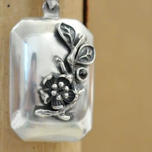 May include: A rectangular silver pendant with a floral design. The pendant features a raised flower, leaves, and a small dark stone. The pendant is suspended from a silver chain. The background is a neutral tan color.
