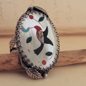 May include: Sterling silver ring with an oval mother-of-pearl inlay, featuring a hummingbird design. The bird is depicted with red, black, and iridescent colours, surrounded by green leaves and red berries. The ring has a detailed, ornate border.