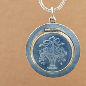 May include: A round, blue and silver pendant necklace. The pendant features a detailed floral basket design in a lighter shade of blue. The necklace has a silver chain.
