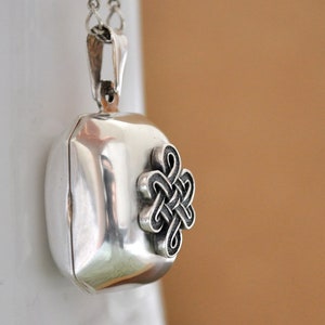 May include: A polished silver locket pendant with a rectangular shape and rounded edges, featuring a raised, intricate, dark-toned Celtic knot design on the front. The locket hangs from a silver chain attached to a sturdy, polished silver bail.