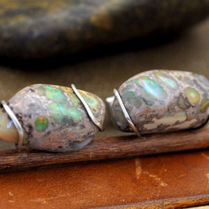 May include: A pair of cufflinks featuring natural stone cabochons with a mix of colors including green, brown, and cream. The stones are wrapped with silver-colored wire and attached to a silver-colored cufflink base. The cufflinks are displayed on a brown surface.