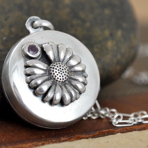 May include: A silver-colored pendant necklace with a daisy design. The round pendant features a raised daisy with a small purple gemstone. The necklace chain is also silver. The pendant is approximately 1 inch in diameter.