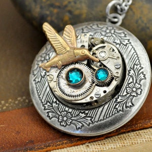 May include: A silver-toned pendant necklace with a steampunk design. The round pendant features a hummingbird, watch gears, and two teal-colored gems. The pendant is approximately 2 inches in diameter and hangs from a silver chain.