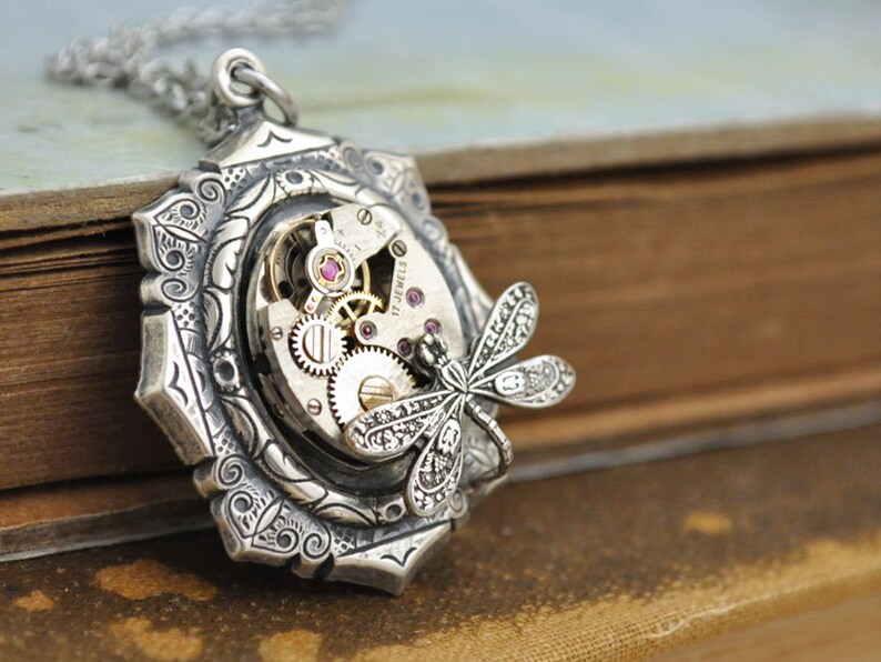 May include: Silver pendant necklace with a detailed floral design and a dragonfly charm. The pendant features a working watch movement with visible gears and a ruby jewel.