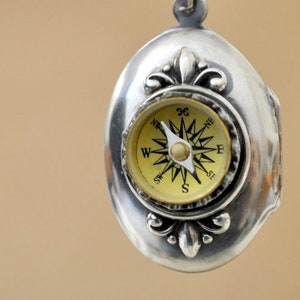 May include: Silver locket with a compass design. The compass face is gold with black markings and a white needle pointing north. The locket has a fleur-de-lis design on the top and bottom.