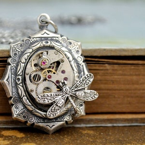 May include: Silver pendant necklace with a detailed dragonfly design and a vintage watch face with exposed gears and ruby accents. The pendant is 17 jewels.