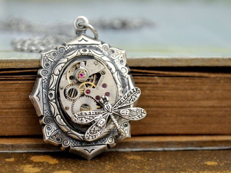 May include: Silver pendant necklace with a detailed dragonfly design and a vintage watch face with exposed gears and ruby accents. The pendant is 17 jewels.