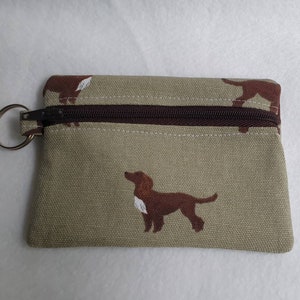 May include: A small, rectangular coin purse with a brown zipper and a silver key ring. The beige fabric is patterned with brown dog silhouettes. The zipper and the trim are brown.