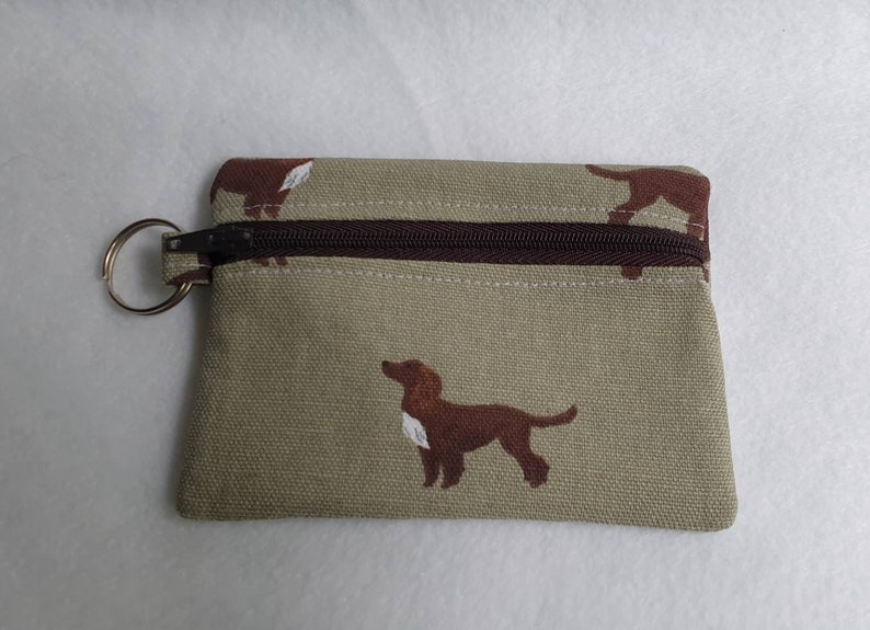 May include: A small, rectangular coin purse with a brown zipper and a silver key ring. The beige fabric is patterned with brown dog silhouettes. The zipper and the trim are brown.