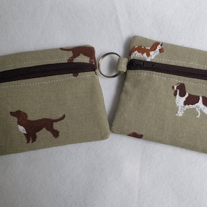 May include: Two small, rectangular coin purses with a brown zipper and a key ring. The purses are beige with a repeating pattern of brown and white dogs. The zipper is brown.