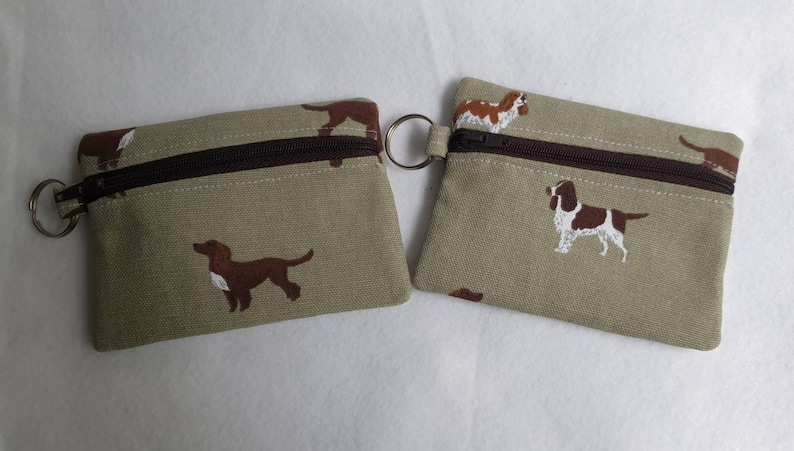 May include: Two small, rectangular coin purses with a brown zipper and a key ring. The purses are beige with a repeating pattern of brown and white dogs. The zipper is brown.