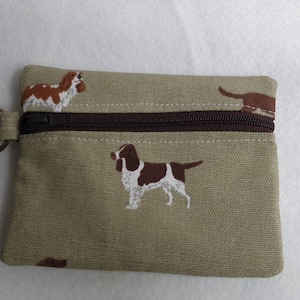 May include: A small, rectangular coin purse in a tan fabric, featuring embroidered brown and white dogs. The purse has a brown zipper closure and a metal key ring attached to the side. The design includes multiple dog illustrations.
