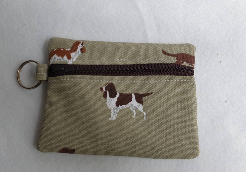 May include: A small, rectangular coin purse in a tan fabric, featuring embroidered brown and white dogs. The purse has a brown zipper closure and a metal key ring attached to the side. The design includes multiple dog illustrations.