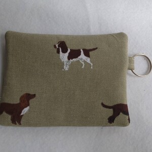 May include: A small, rectangular, olive-green fabric pouch with a key ring attached. The pouch is decorated with brown and white dog illustrations. The dogs are in various poses. The pouch is likely used to hold keys or small items.