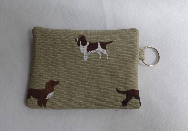 May include: A small, rectangular, olive-green fabric pouch with a key ring attached. The pouch is decorated with brown and white dog illustrations. The dogs are in various poses. The pouch is likely used to hold keys or small items.