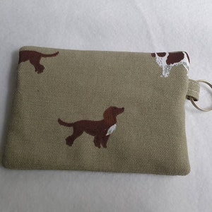 May include: A small, rectangular coin purse in a tan fabric, adorned with repeating brown and white dog illustrations. The purse has a small loop with a silver key ring attached.