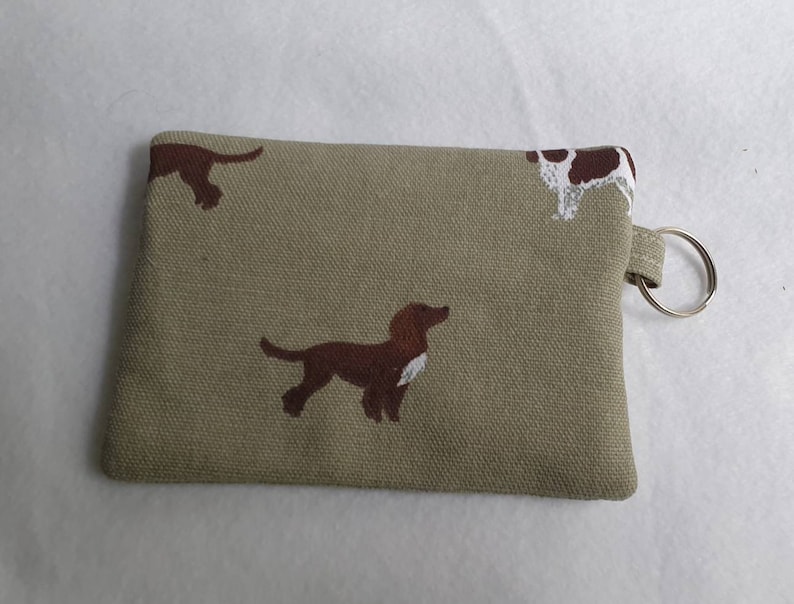 May include: A small, rectangular coin purse in a tan fabric, adorned with repeating brown and white dog illustrations. The purse has a small loop with a silver key ring attached.