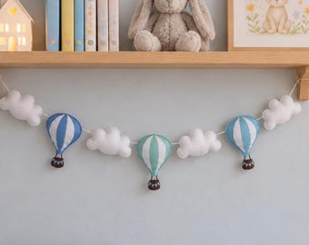 May include: A decorative garland featuring three fabric hot air balloons in blue, white, and mint green, interspersed with four white plush clouds. The balloons have brown baskets and are strung together on a white cord for hanging display.