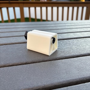 May include: A small, off-white, rectangular object with a black cylindrical component on one end. The object is resting on a gray, textured surface, likely a table. The background is blurred, showing a wooden deck and foliage.