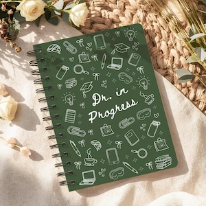 May include: A spiral-bound notebook with a dark green cover featuring white illustrations of school and office supplies. The words "Dr. in Progress" are written in white script. The notebook is surrounded by flowers.