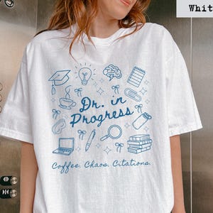 May include: White t-shirt with a blue graphic design. The design includes a graduation cap, lightbulb, brain, notepad, coffee cup, and the text "Dr. in Progress." Below the text is "Coffee. Chaos. Citations."
