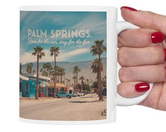 Op de afbeelding: Een witte keramische mok met een kleurrijk beeld van palmbomen en een retro-ontwerp. De tekst op de mok luidt "Palm Springs. Come for the sun, stay for the fun."