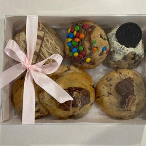 May include: A white box with a pink ribbon tied around it. The box contains six cookies, each with a different topping. The toppings include chocolate chips, sprinkles, and a chocolate bar.