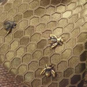 Puede incluir: Un bolso de color dorado con un patrón de panal y tres abejas como adorno.
