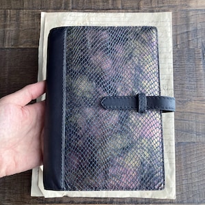 May include: A rectangular notebook cover featuring a textured snakeskin pattern in shades of black, purple, and gold. The cover includes a solid black leather spine and a matching black leather strap closure that secures the front panel horizontally.