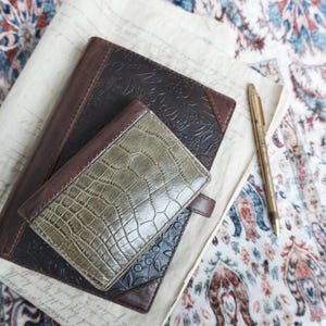 May include: A dark brown leather journal with an embossed floral pattern, a smaller olive green crocodile-textured leather wallet placed on top, and a gold-toned metal ballpoint pen resting beside them on a piece of off-white parchment paper.