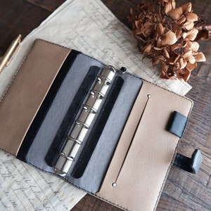 May include: A brown leather bound planner with a silver ring binder system. The planner is open and has a gray and black interior. The planner is on a wooden surface.