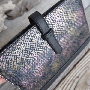 May include: A rectangular wallet featuring a metallic, iridescent snakeskin pattern in shades of purple, gold, and black. The wallet is secured with a solid black leather strap and a matching black trim along the edges, creating a structured look.