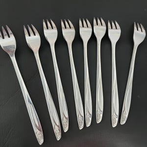May include: A set of eight stainless steel forks with a three-tine design. The forks have a slender handle with a decorative floral pattern near the base. The forks are arranged on a black surface.