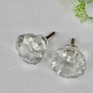 May include: Two clear, faceted glass cabinet knobs with a multi-sided, geometric shape. Each knob features a central metal screw post with a brass-toned finish, designed for attachment to furniture drawers or cabinet doors for functional use.