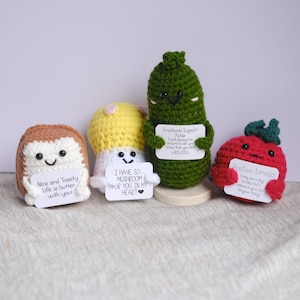 Puede incluir: Cuatro peluches de comida tejidos a crochet que sostienen carteles con mensajes alentadores. Un peluche de pan marrón dice "Nice and Toasty Life is butter with you!" Un peluche de hongo blanco dice "I have so... mushroom for you in my heart." Un peluche de pepinillo verde dice "Emotional Support Pickle I will always be around to let you know that you are a BIG dill!" Un peluche de tomate rojo dice "Positive tomato I may be a tiny tomato but I believe in you! Go do your thing!"