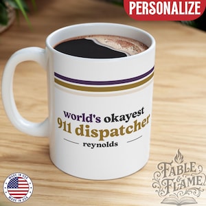 Puede incluir: Taza de café de cerámica blanca con asa, llena de café oscuro. La taza presenta el texto "world's okayest 911 dispatcher" en una fuente estilizada, con el nombre "reynolds" debajo. La taza tiene rayas moradas y doradas.