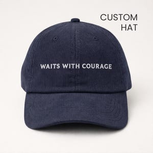 Puede incluir: Gorra de béisbol de pana azul marino con el texto blanco "WAITS WITH COURAGE". Las palabras "CUSTOM HAT" están impresas encima. La gorra tiene una visera curva y una parte superior redondeada.