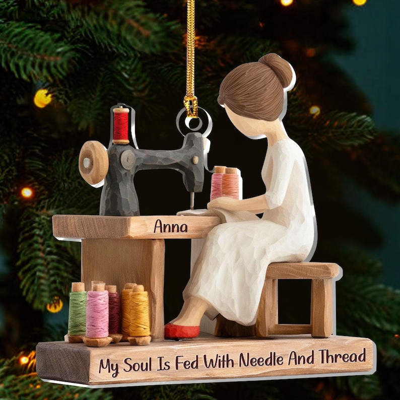 May include: A wooden Christmas ornament depicting a person sewing at a vintage sewing machine. The ornament features a person in a white dress, a sewing machine, spools of thread, and the text "My Soul Is Fed With Needle And Thread". The name "Anna" is also visible.