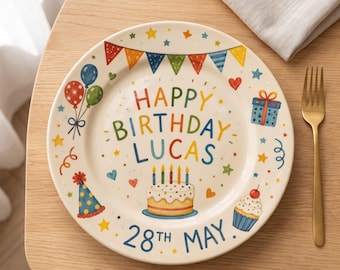 May include: A round ceramic plate featuring a colorful birthday illustration with the text HAPPY BIRTHDAY LUCAS and 28TH MAY. The design includes a cake with five candles, a cupcake, balloons, a party hat, a gift box, and bunting flags.