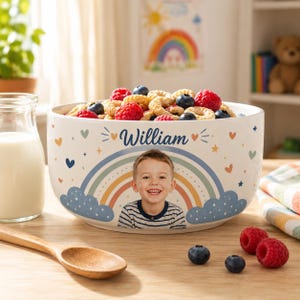 Può includere: Una ciotola in ceramica bianca riempita di cereali, decorata con un arcobaleno, nuvole blu, piccoli cuori, stelle e il nome William in blu. Una fotografia di un bambino piccolo è centrata sulla parte anteriore della ciotola sotto il nome.